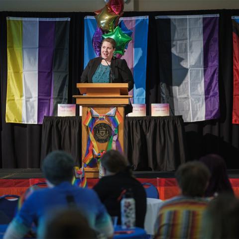 Woman standing at podium with pride flags in background.